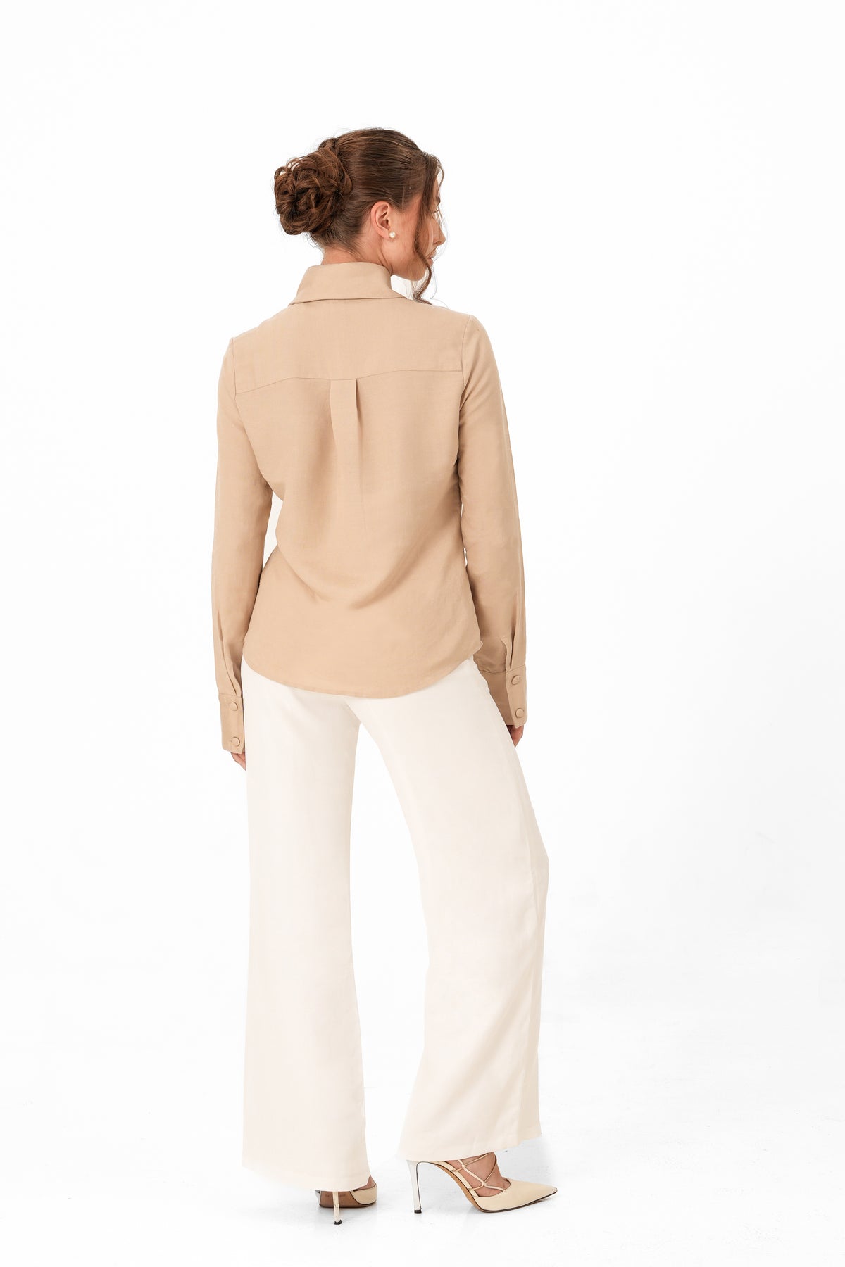 A woman is shown from the back wearing a beige long-sleeve collared shirt and off-white wide-leg trousers, paired with beige high heels, against a plain white studio background.