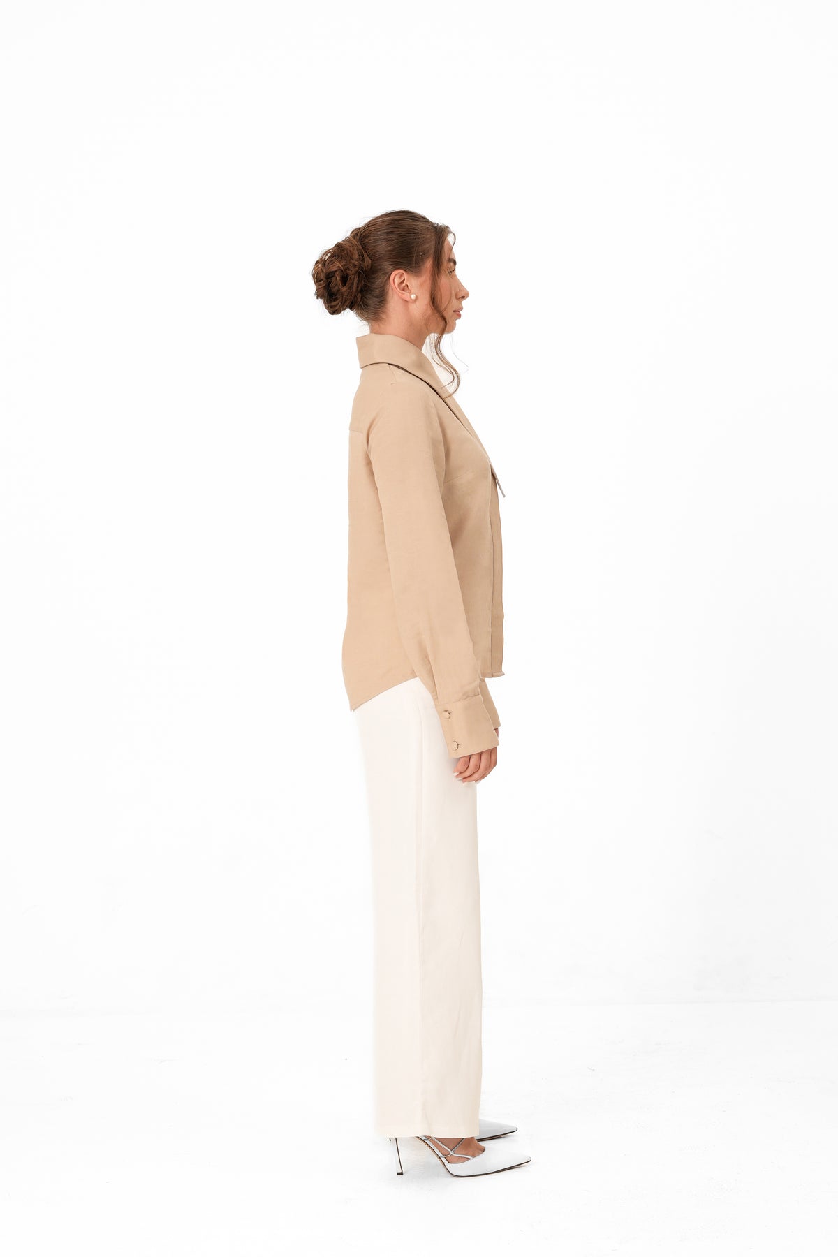 A woman shown in profile wearing a beige long-sleeve button-front top, white wide-leg pants, and white pointed heels against a clean white studio background.