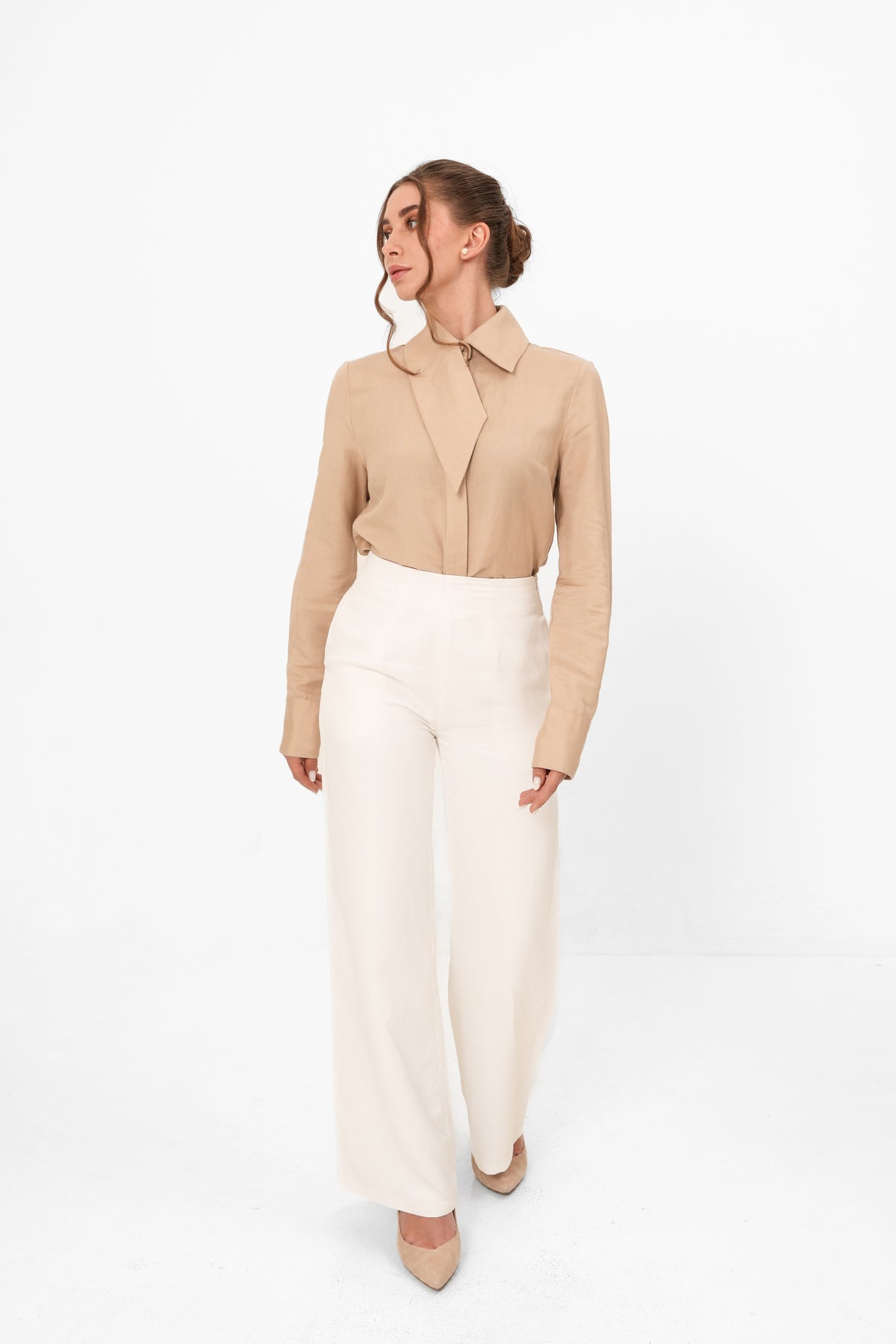 A woman modeling a beige long-sleeve collared blouse tucked into high-waisted, wide-leg cream trousers, photographed against a seamless white studio background.