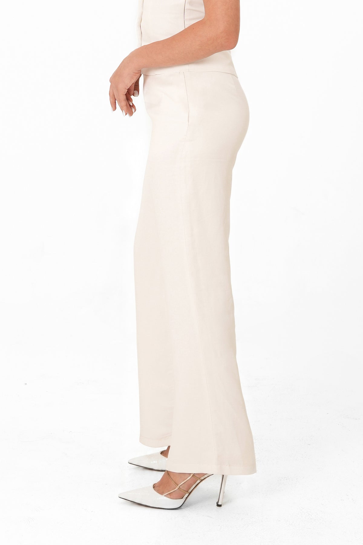 A model shown from the side wearing a high-waisted, off-white maxi skirt with a slight flare at the hem, paired with white pointed-toe heels on a white studio background.