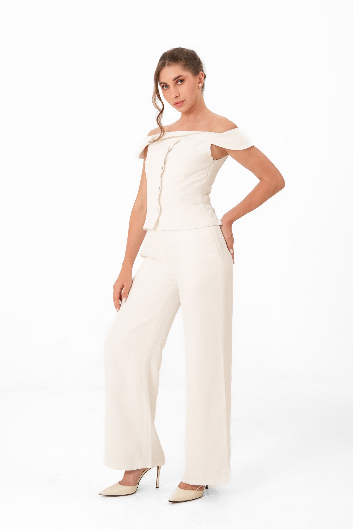 A woman modeling an off-the-shoulder cream jumpsuit with a structured bodice and wide-leg trousers, paired with beige heels, against a plain white studio background.