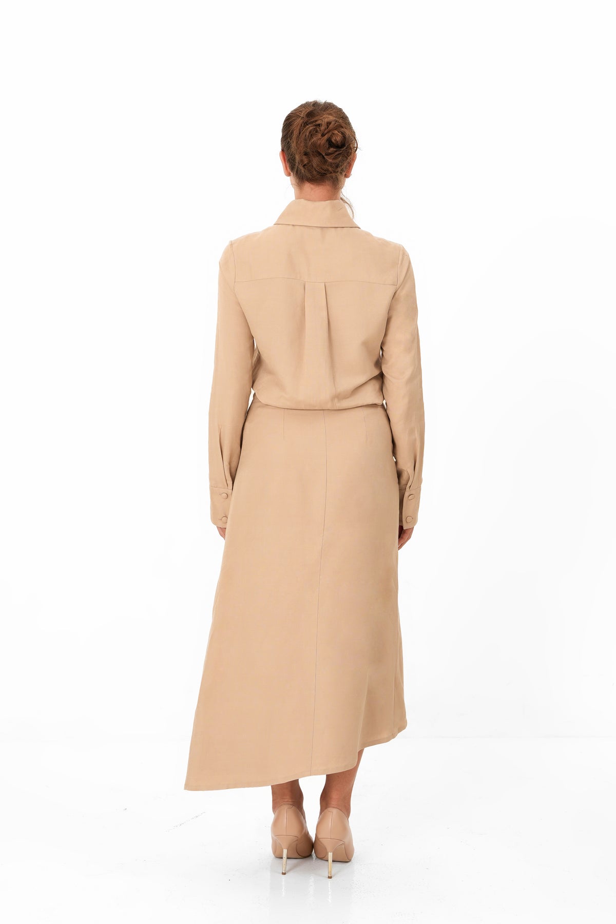 A woman viewed from behind wearing a long beige trench coat with a matching belt and high collar, paired with beige heels, against a plain white studio background.