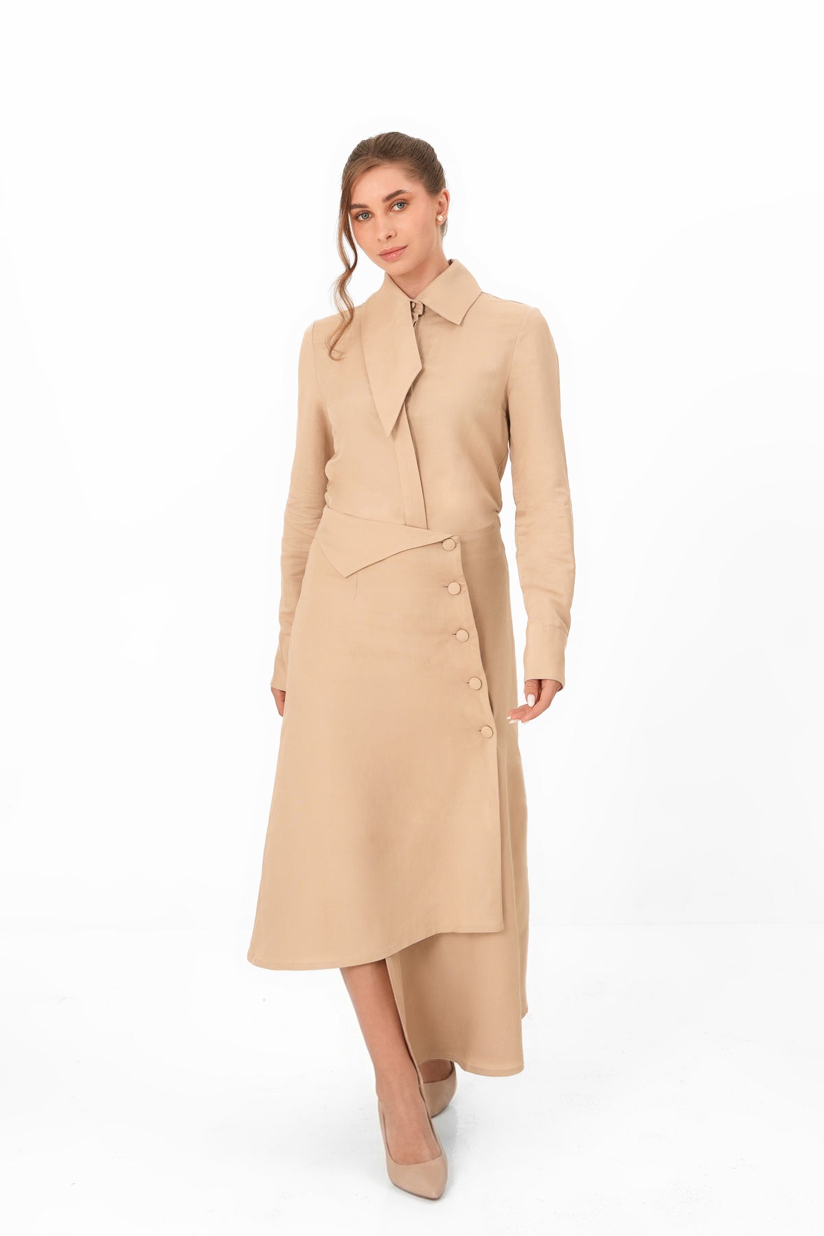 A woman modeling a long-sleeve beige wrap-style midi dress with a high-low hem and front buttons, photographed against a clean white studio background.