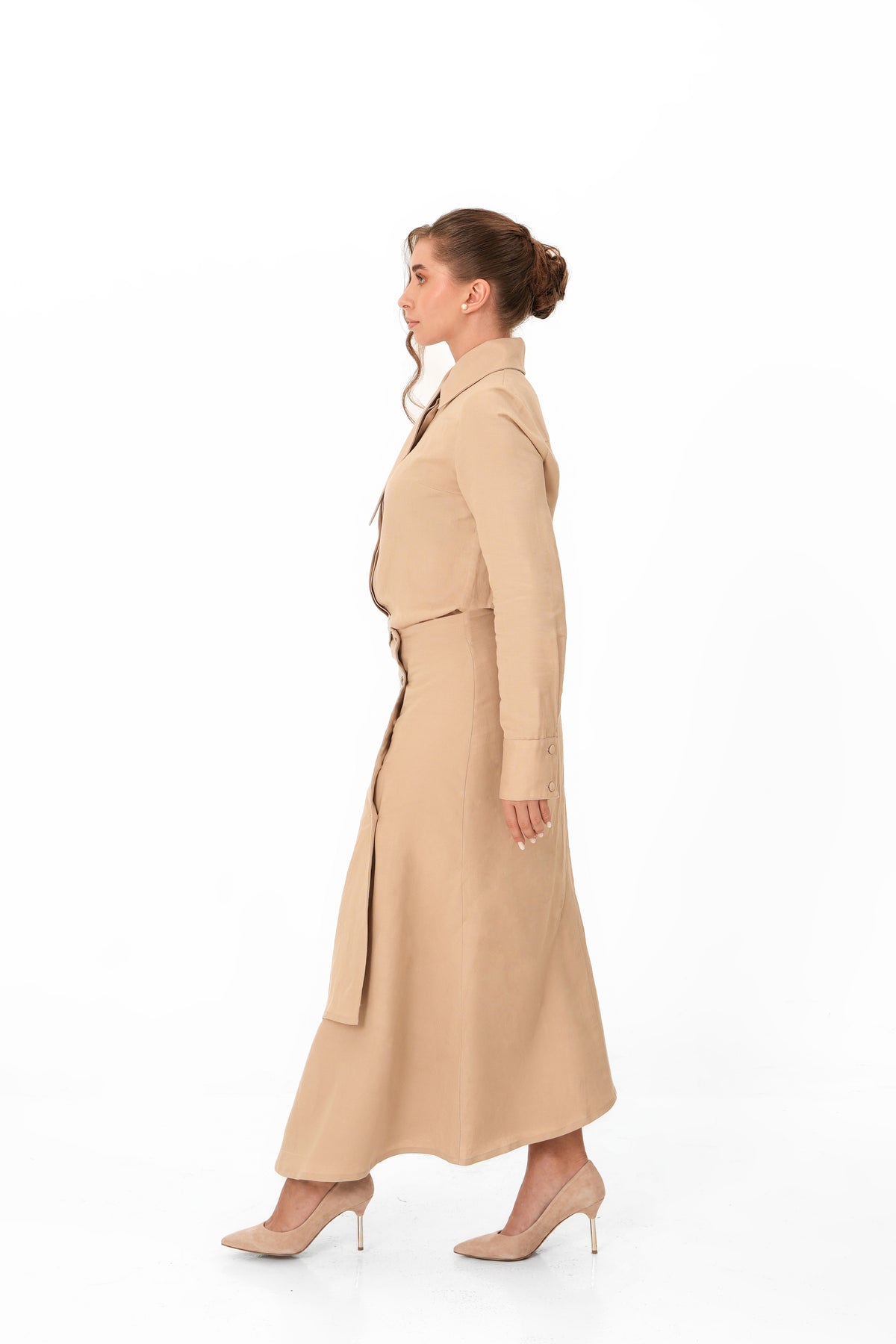 A woman in profile wearing a long beige trench-style dress with a matching belt and nude high heels, photographed against a clean white studio background.