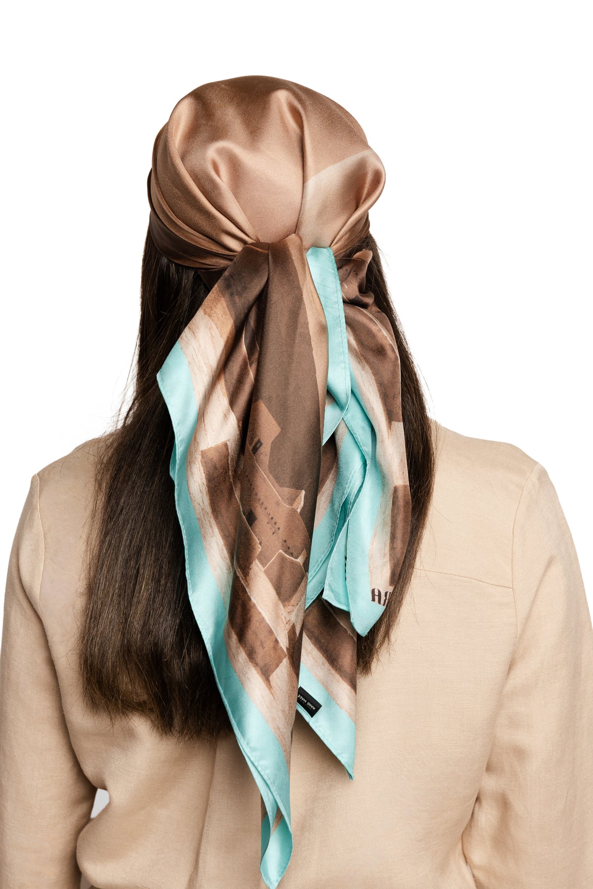 A woman shown from the back wearing a brown satin scarf tied around her head and draped over her shoulders. The scarf has a geometric pattern in brown, beige, and light blue.