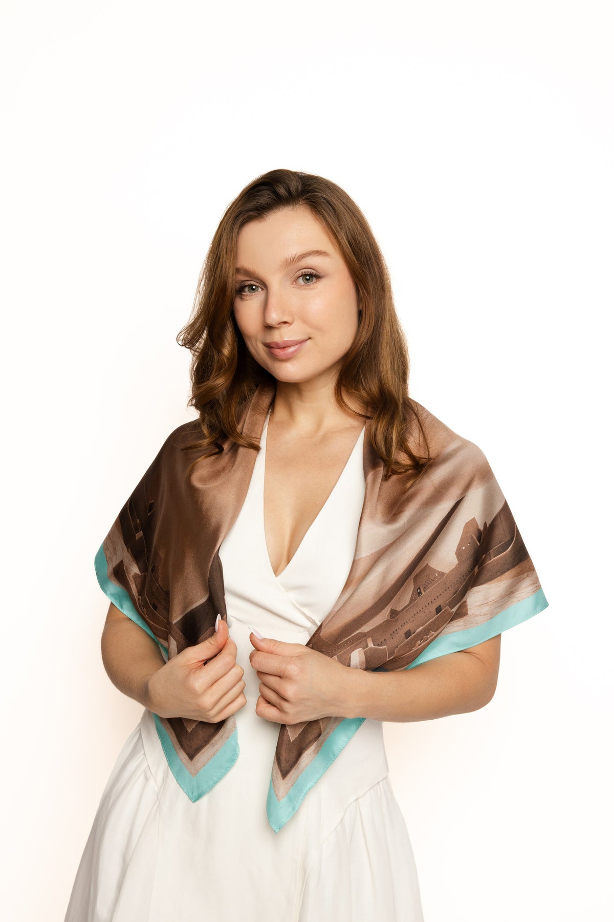 A woman with light brown hair is wearing a white dress and draping a square silk scarf with a brown and beige landscape print and turquoise blue borders, set against a plain white background.