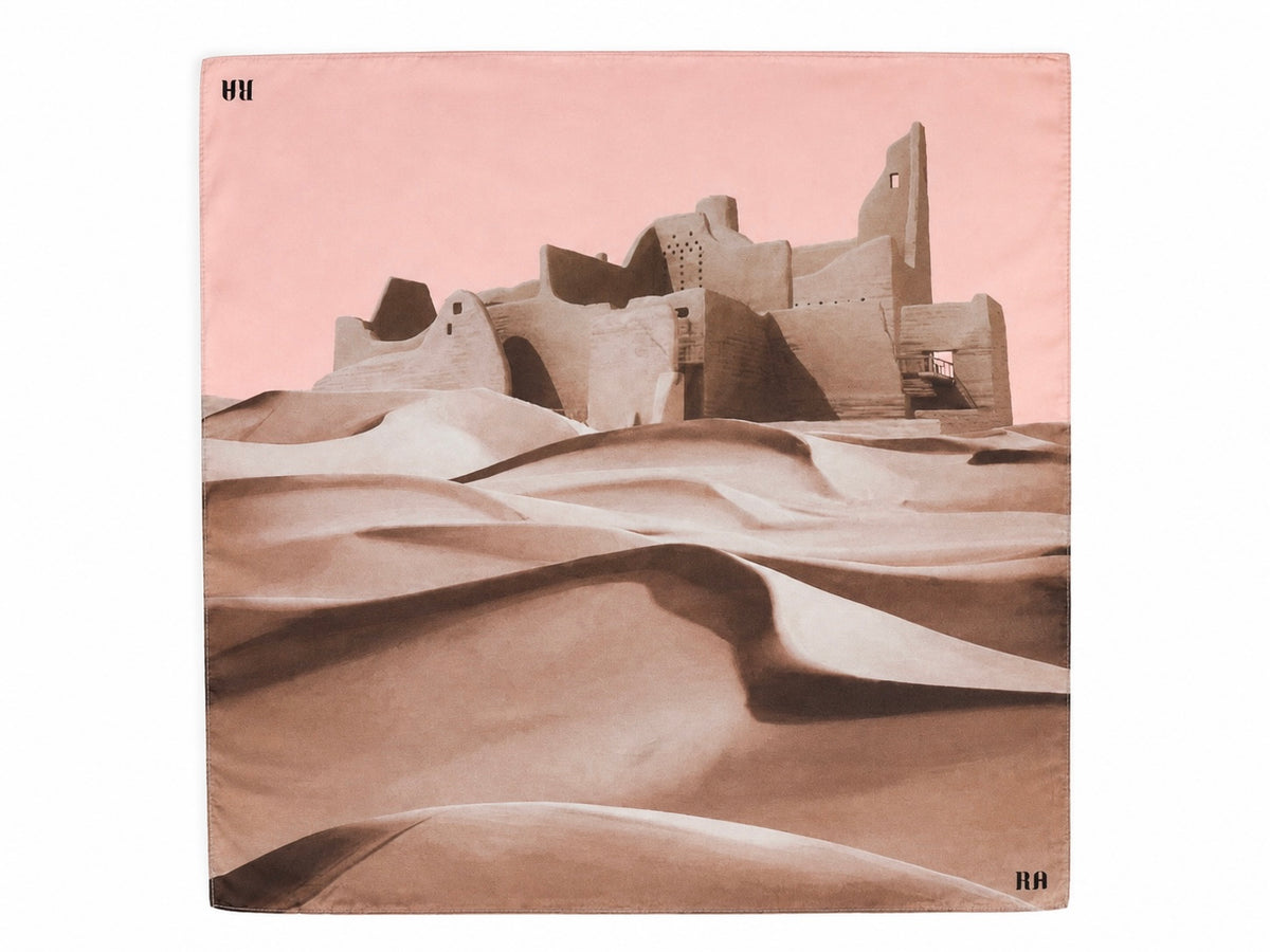 A square scarf featuring a desert landscape print with sand dunes and a historic adobe-style fortress under a dusty pink sky. The scarf has a white border with small black 'HA' monogram logos in the top-left and bottom-right corners.