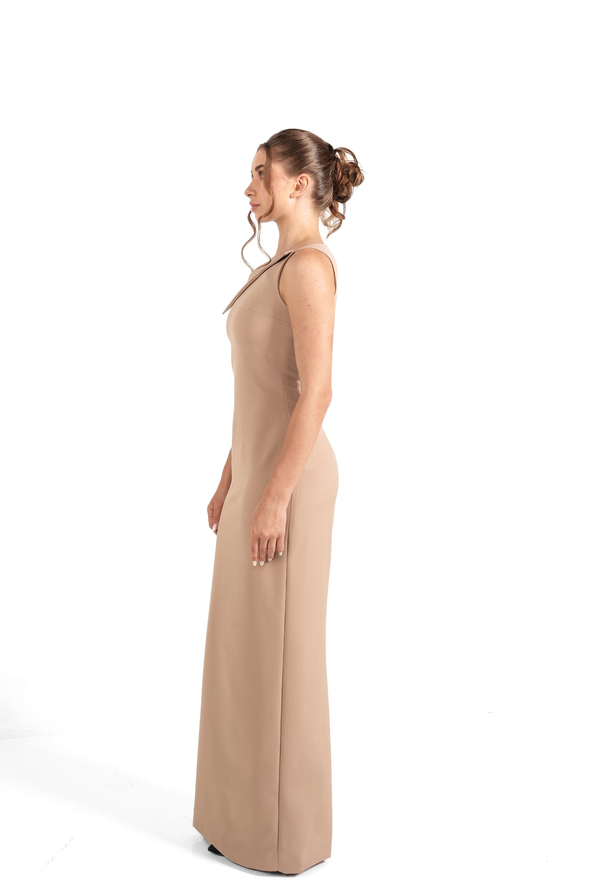 A woman in profile wearing a sleeveless, floor-length beige gown against a plain white studio background.