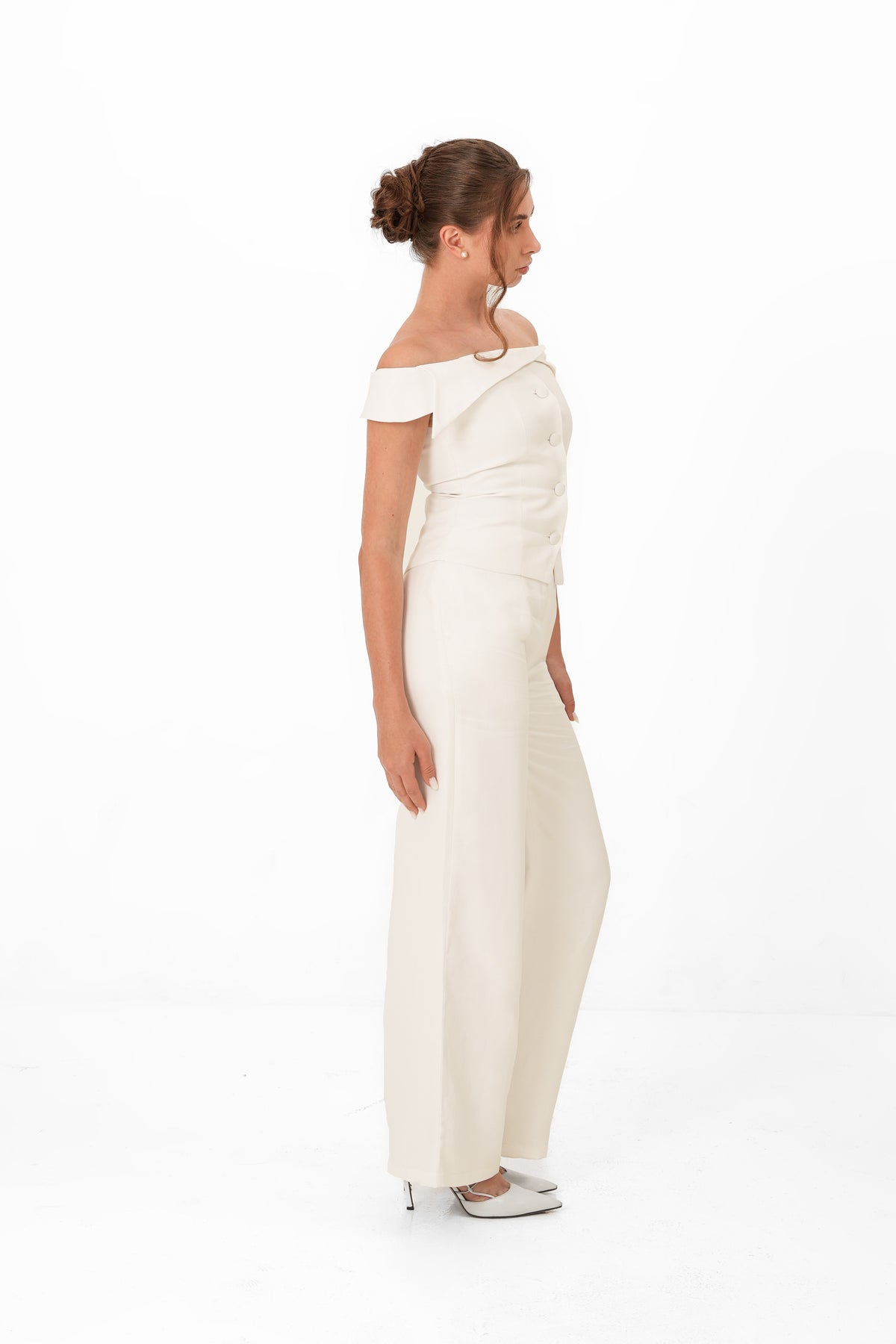 A woman modeling an off-the-shoulder cream-colored maxi dress with a draped neckline and wide-leg pants, shown from a side profile against a plain white studio background.