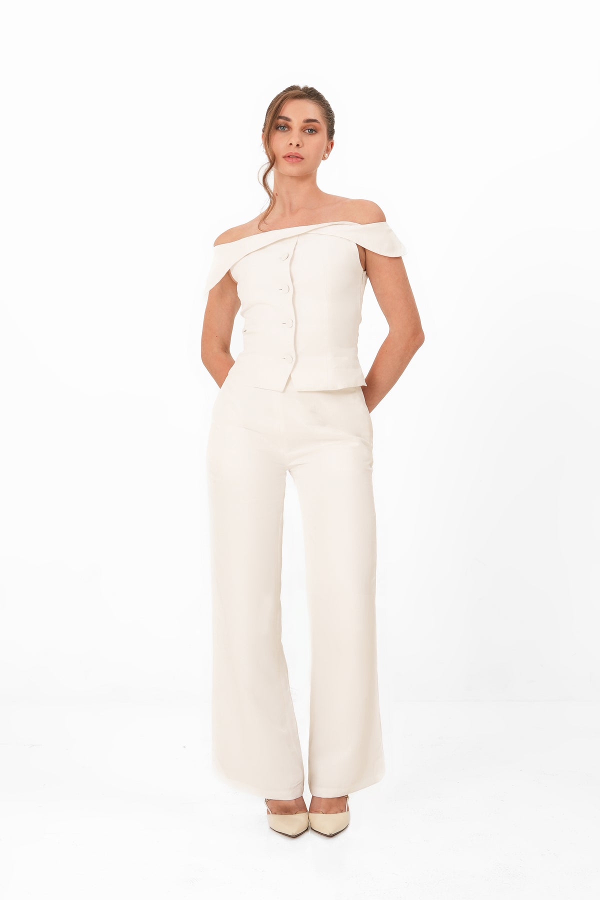 A woman modeling an off-the-shoulder cream-colored jumpsuit with a structured bodice and wide legs, paired with matching pointed-toe heels, against a plain white studio background.