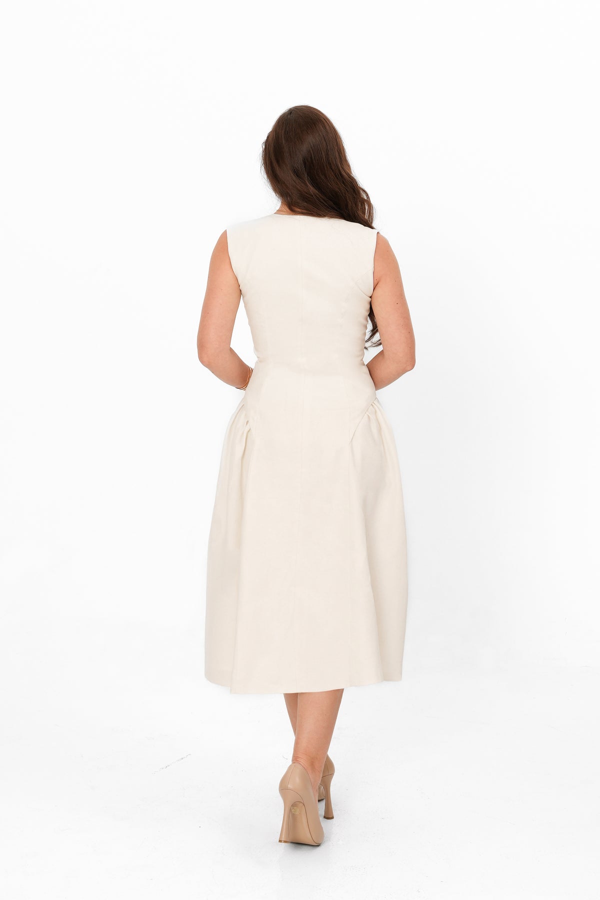 A woman viewed from behind wearing a sleeveless, knee-length cream dress and beige heels against a plain white studio background.