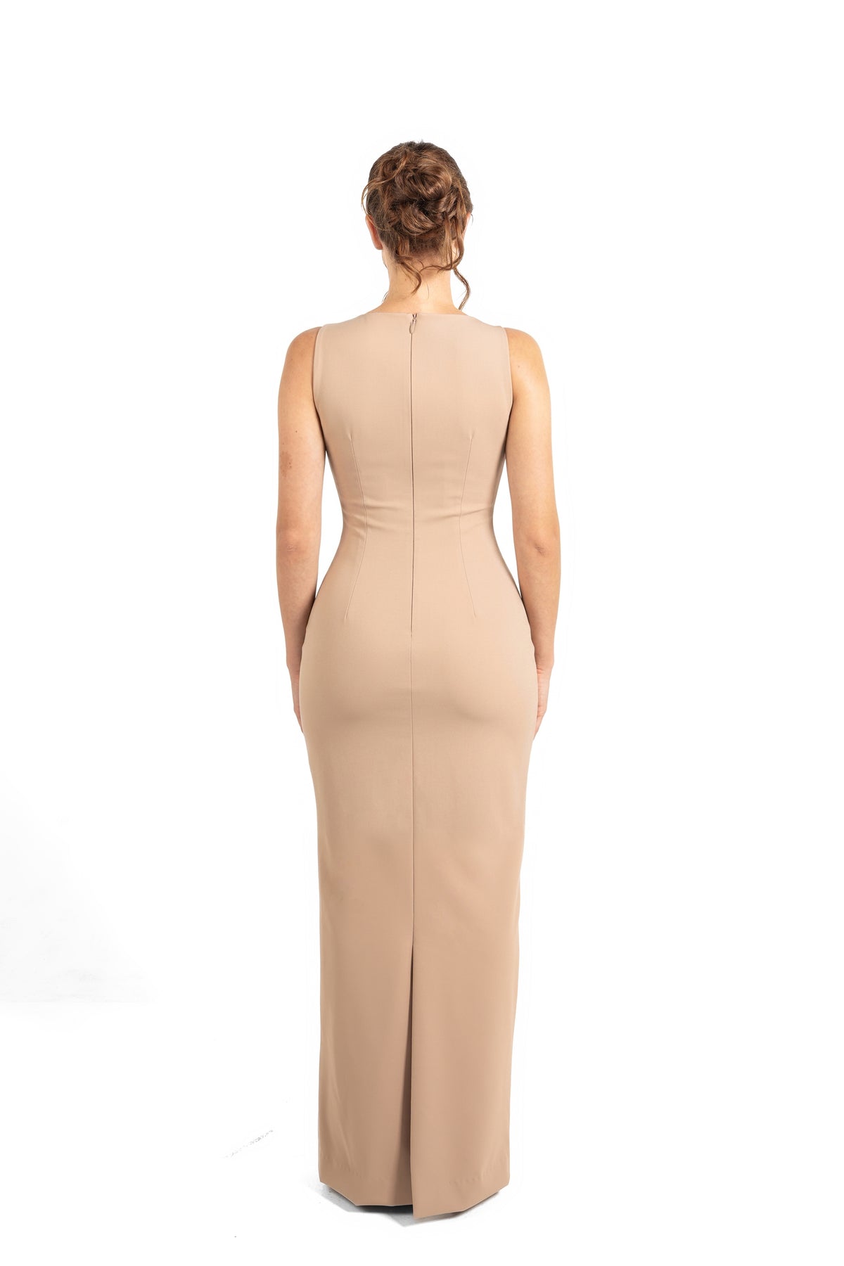 A woman viewed from the back wearing a sleeveless, floor-length beige gown with a fitted silhouette and a front slit, photographed against a plain white studio background.