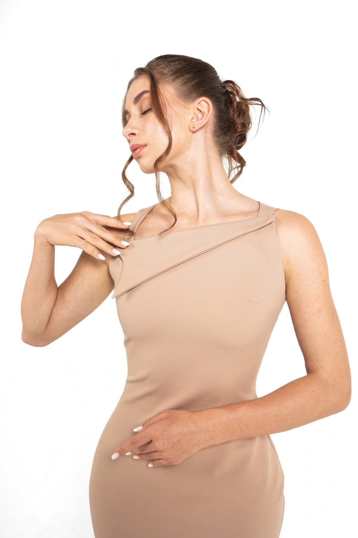 A woman modeling a sleeveless, asymmetrical beige dress against a solid white background.