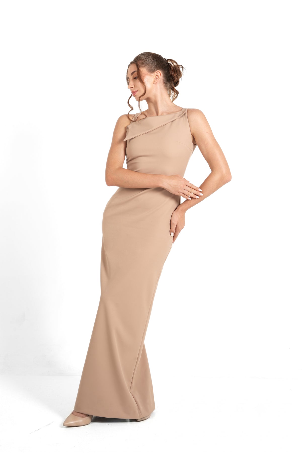A woman modeling a floor-length, one-shoulder beige gown against a plain white studio background.
