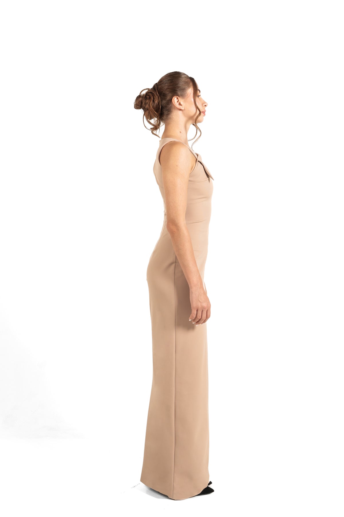 A woman in side profile wearing a sleeveless, floor-length beige gown against a plain white studio background.